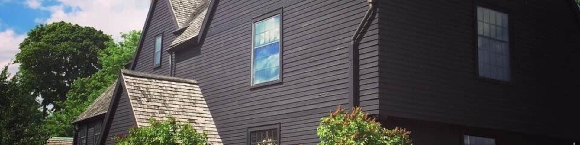 Uma casa de cor escura com vários telhados empenaados e um jardim bem cuidado, com arbustos diversos e uma escultura de pássaro.
