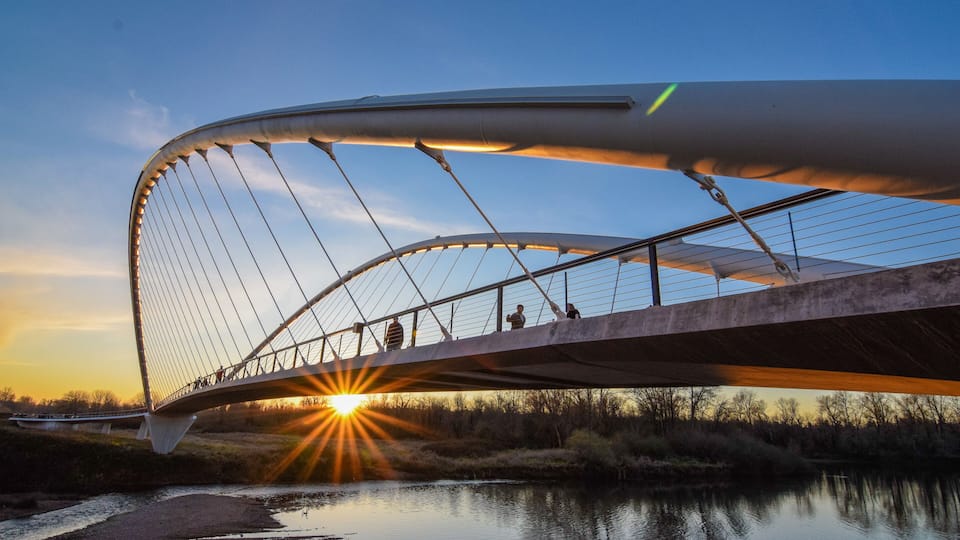 A modern bridge with a curved design over a calm river at sunset.