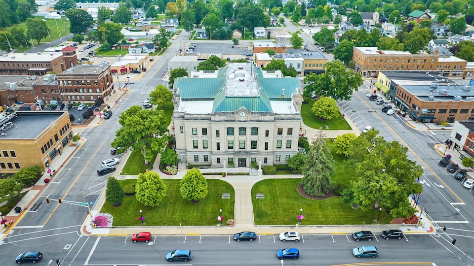 A cityscape with a prominent building with a green roof, surrounded by trees and parked cars.