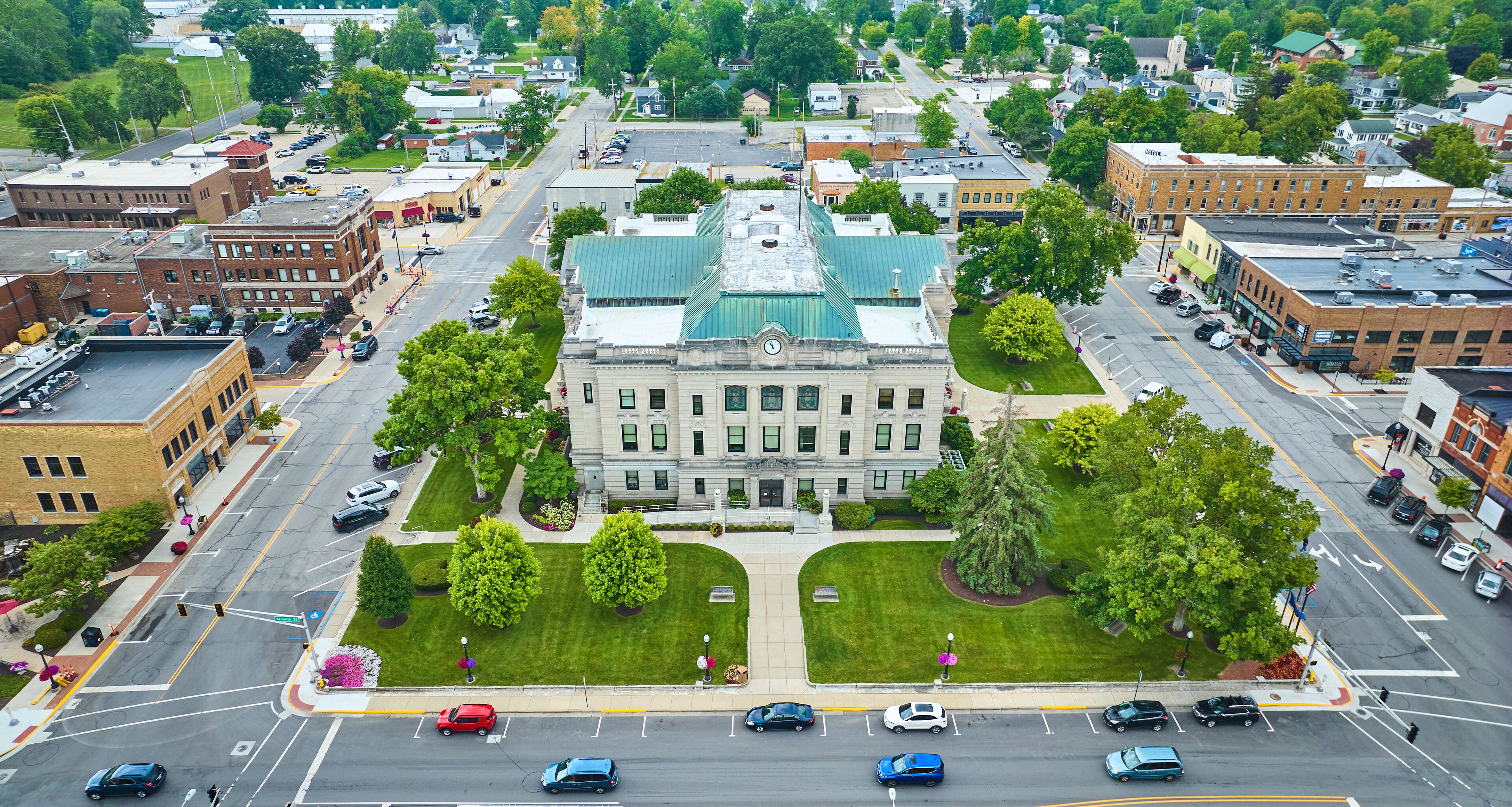 A cityscape with a prominent building with a green roof, surrounded by trees and parked cars.