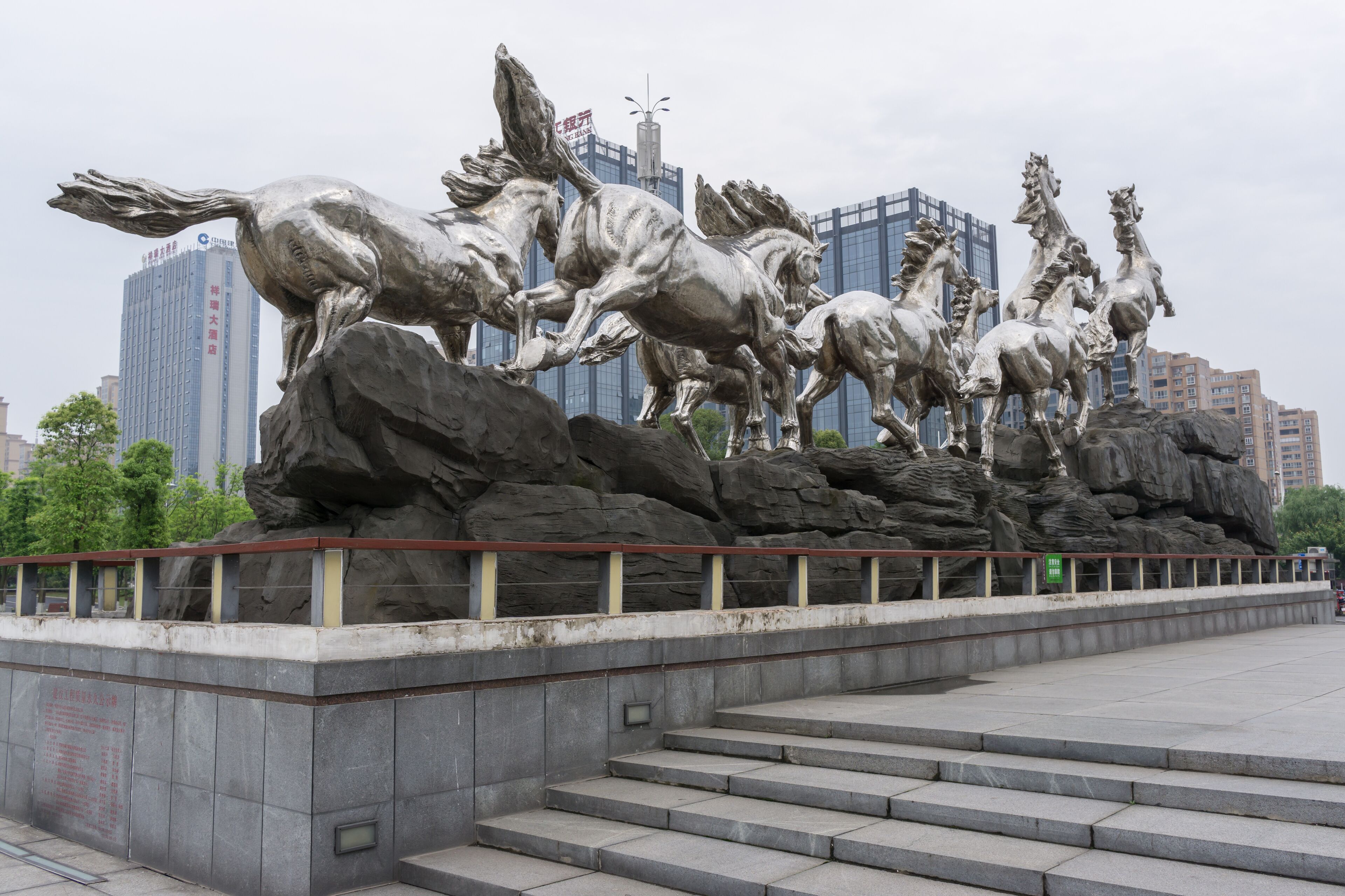 A large metallic sculpture of horses on rocks with a cityscape in the background.