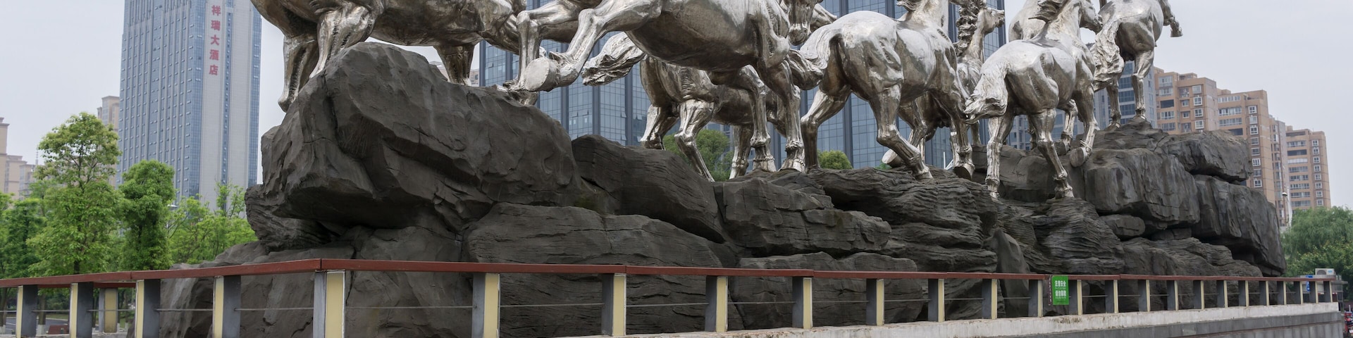 A large metallic sculpture of horses on rocks with a cityscape in the background.