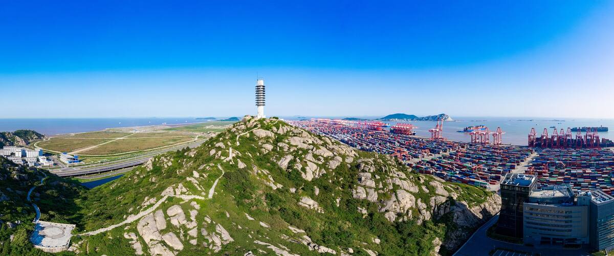 A coastal port with shipping containers, a lighthouse, and a mountainous landscape.