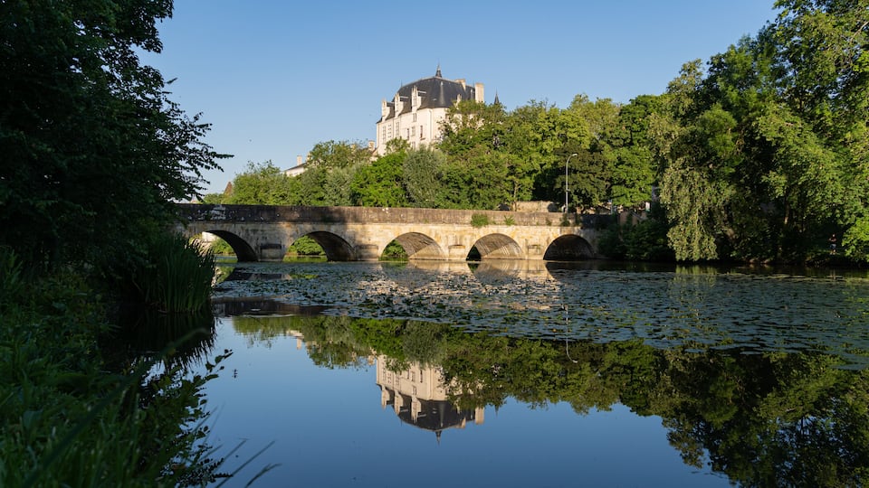 A stone bridge over a calm river with a castle in the background.