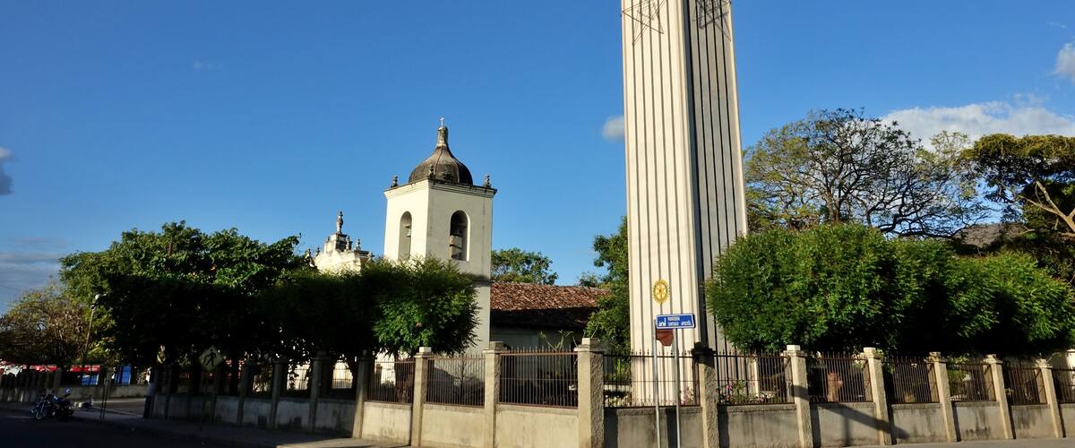 A clock tower with a dome and bell tower in the background, surrounded by trees and a fence.