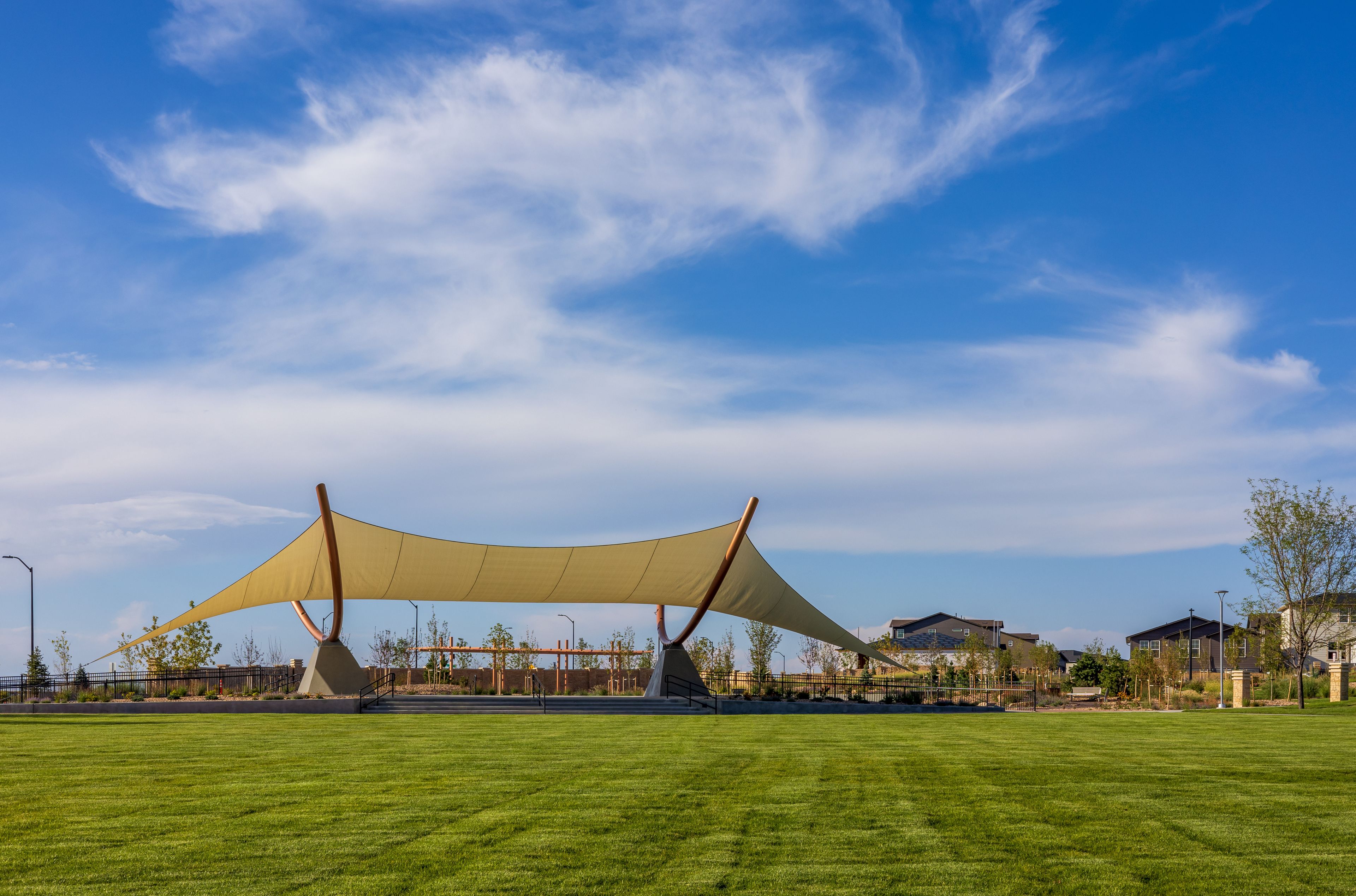 A park with a large shade structure, green grass, and houses in the background.