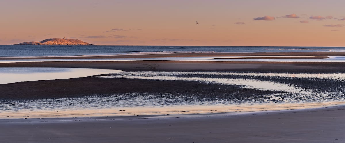 A beach with calm waters, a distant island, and a clear sky.