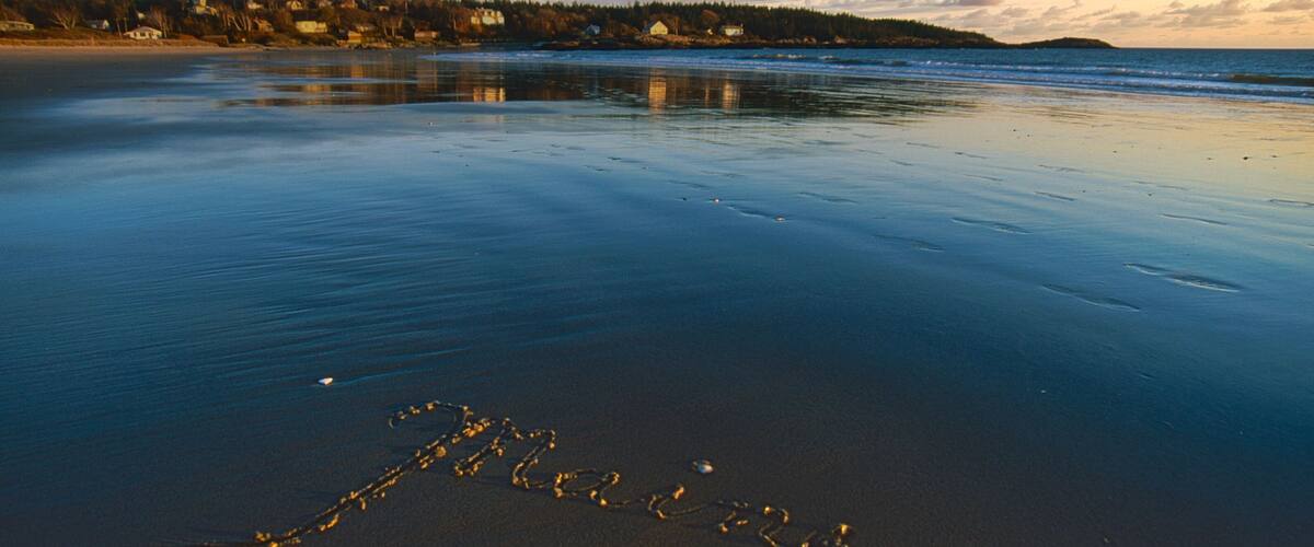 Una playa con la palabra "Maine" escrita en la arena, rodeada de agua tranquila y una costa lejana con casas y árboles.