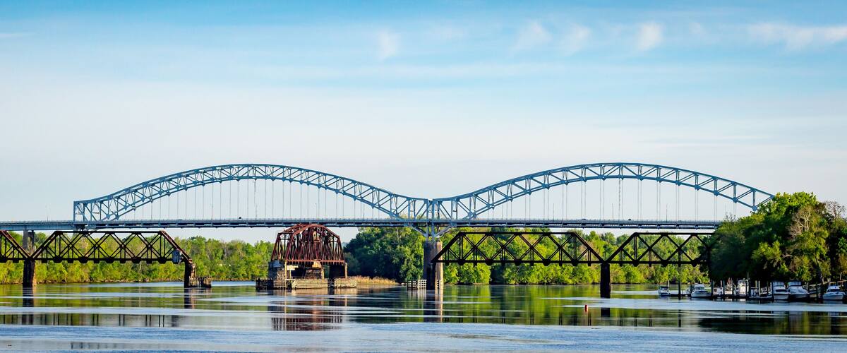 A steel bridge with a truss design spanning over a calm river, surrounded by lush greenery.