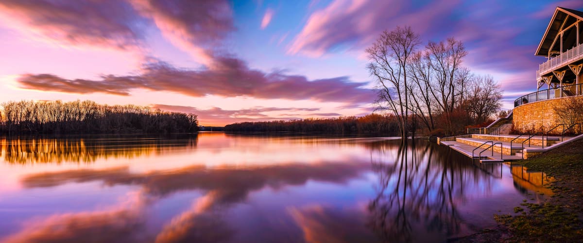 A serene lake with a sunset, trees, and a stone building with stairs.