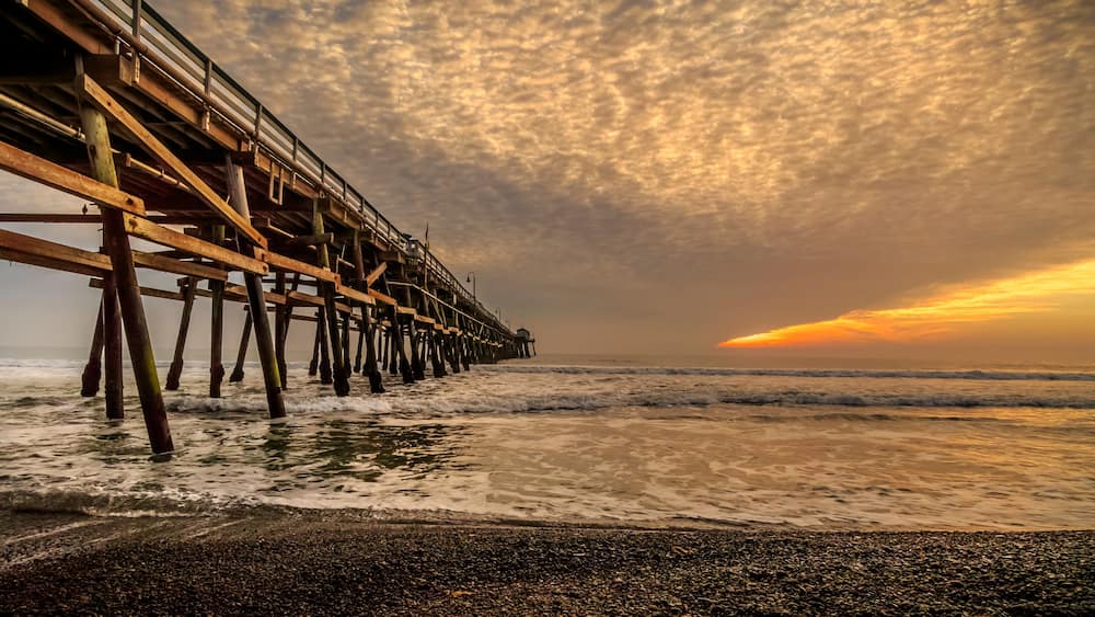 A wooden pier extending into the ocean with waves crashing against it under a cloudy sky.