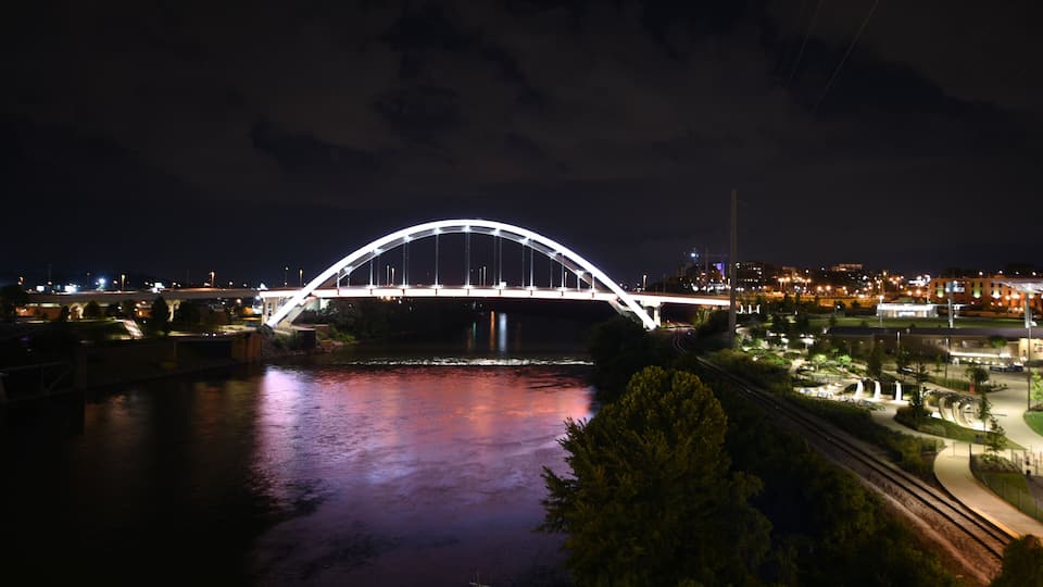 A nighttime view of a large arched bridge over a river, with city lights in the background.