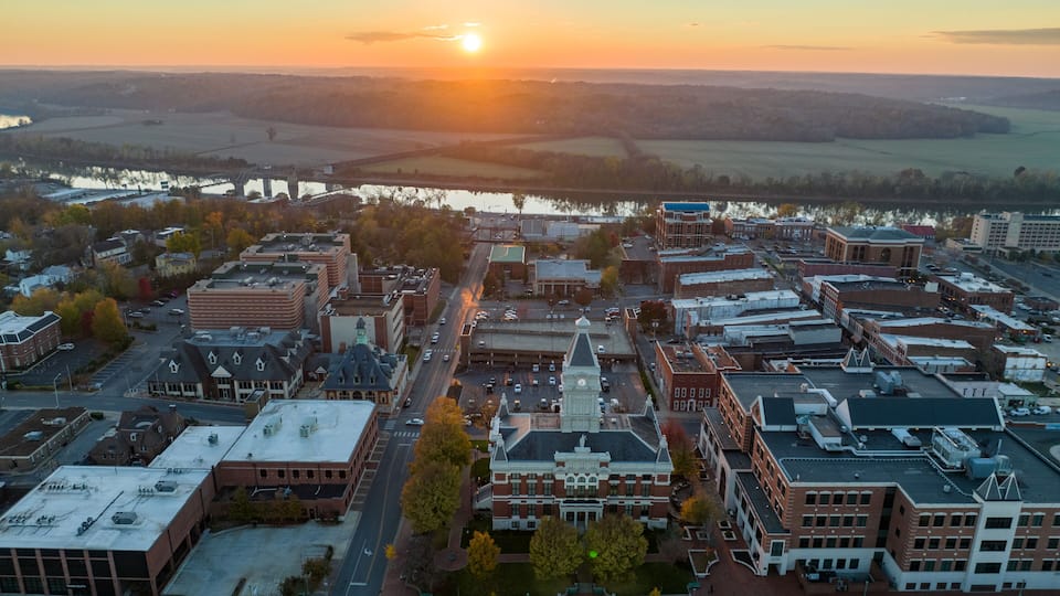 A cityscape at sunset with a prominent clock tower, a river, and surrounding fields.