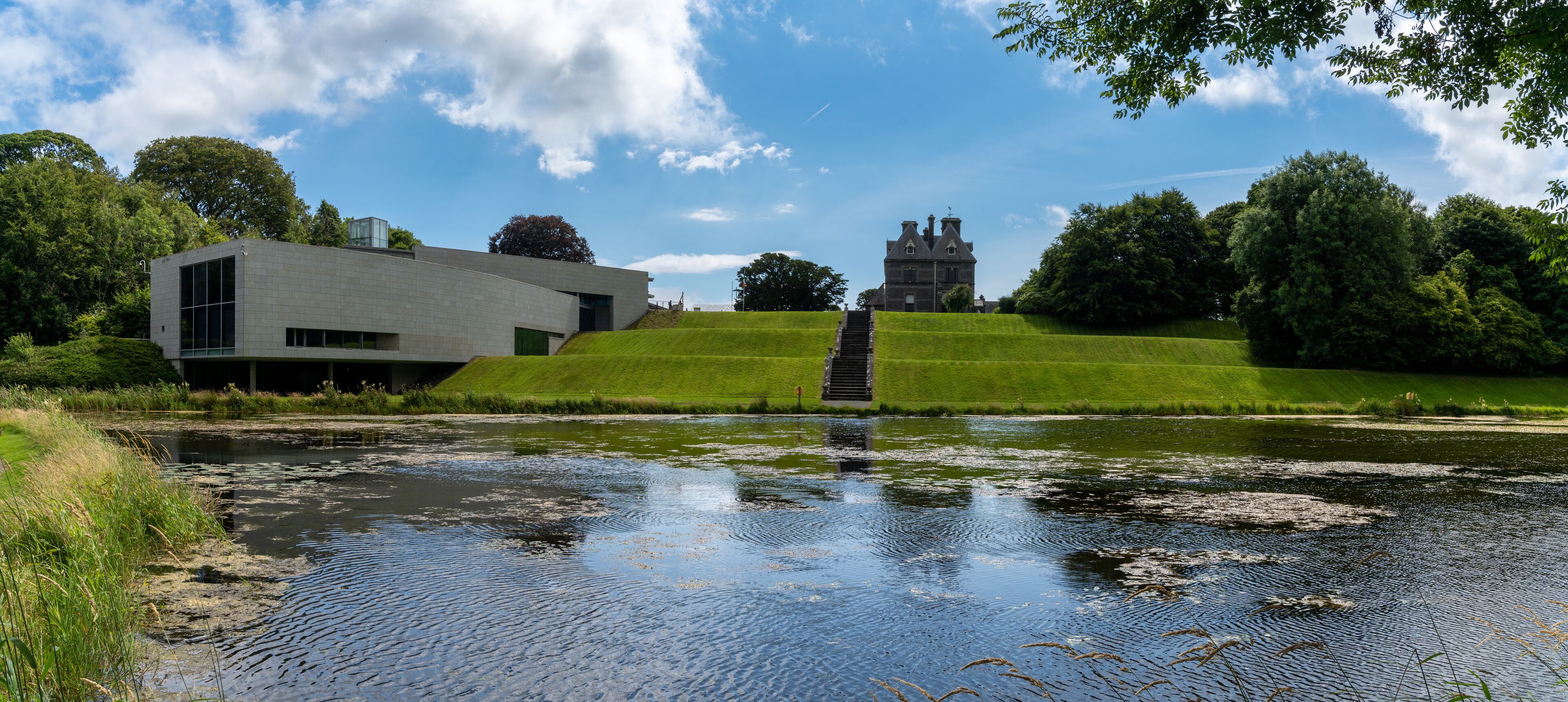A modern building, a traditional house, and a grassy hill with a pond.