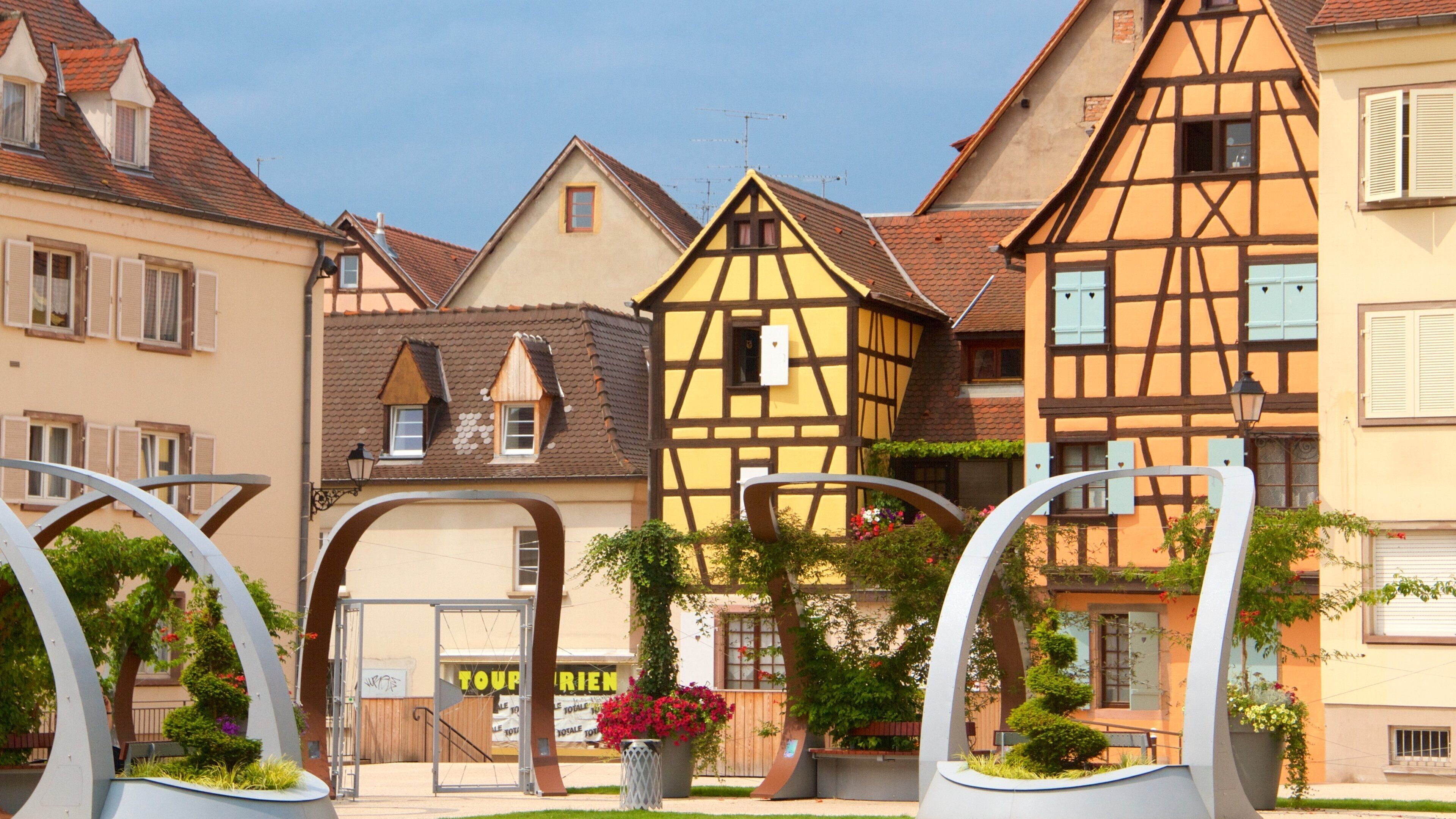 A European town with traditional half-timbered houses, a gate with a sign, and a flower bed.