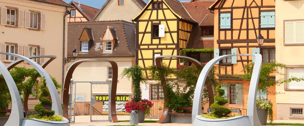 A European town with traditional half-timbered houses, a gate with a sign, and a flower bed.