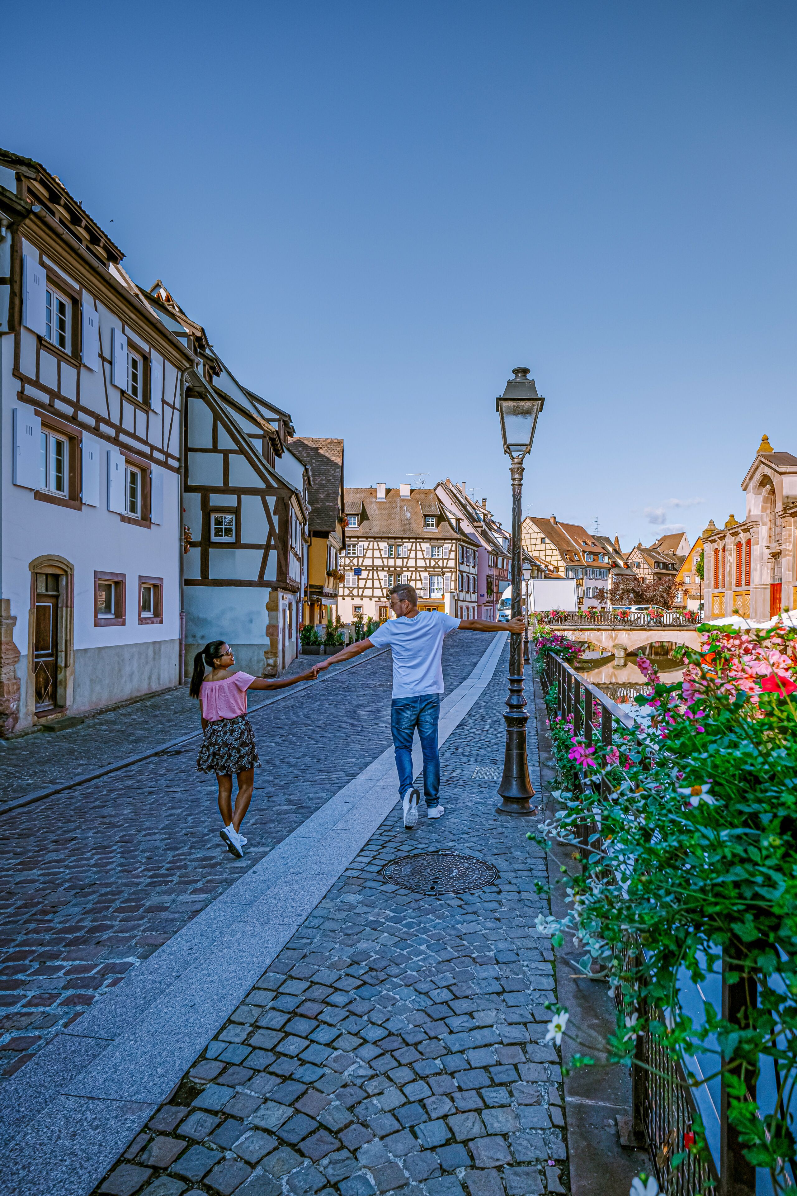A cobblestone street lined with traditional half-timbered buildings, a pedestrian bridge, and a lamppost.