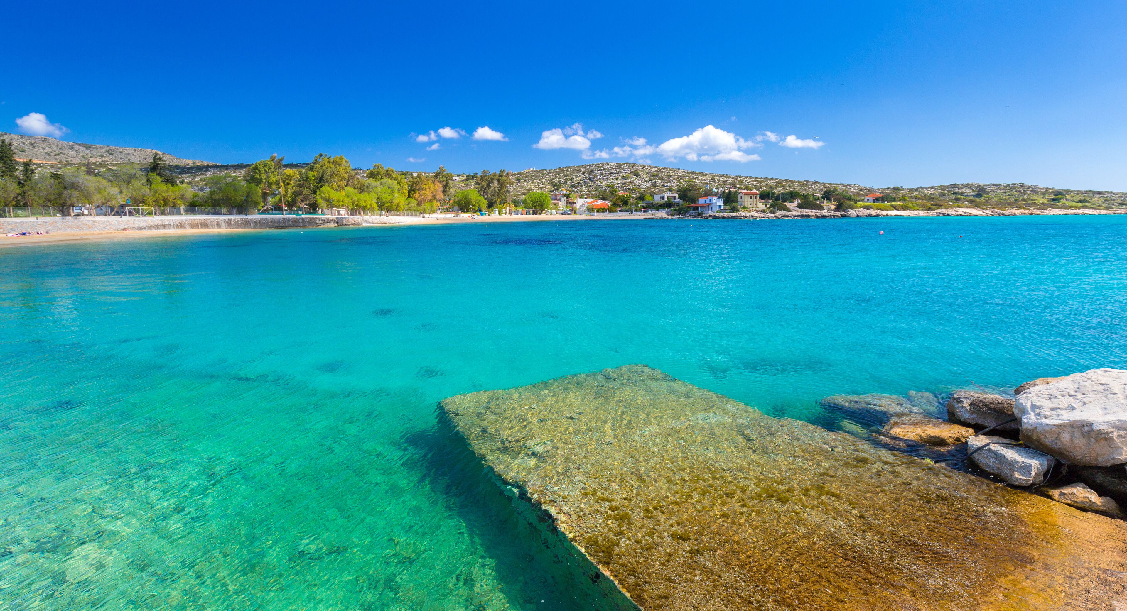 A clear turquoise sea, a rocky outcrop, and a distant shoreline with buildings and trees.