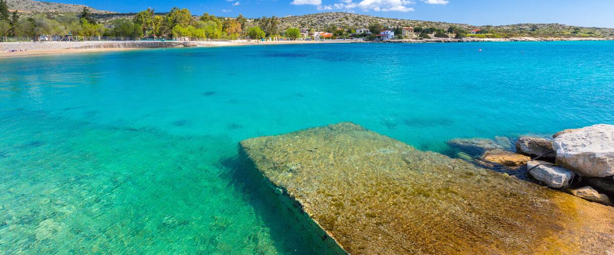 A clear turquoise sea, a rocky outcrop, and a distant shoreline with buildings and trees.