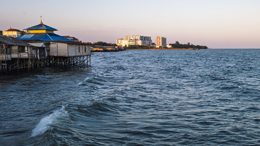 Escena costera con un muelle que se adentra en el mar, un pequeño edificio con techo azul y un paisaje urbano al fondo.