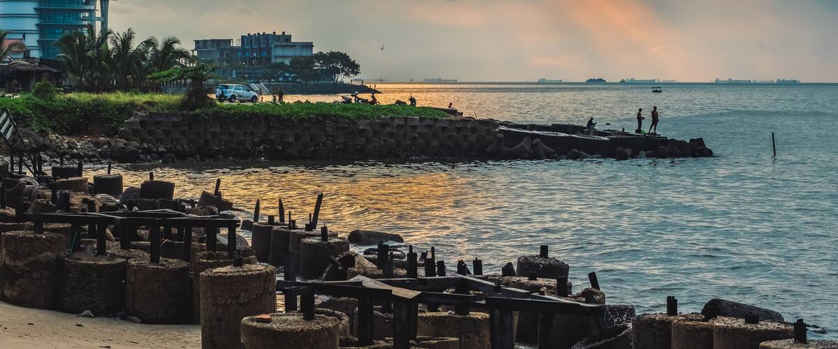 A waterfront with a stone pier, a modern building, and a sunset sky.