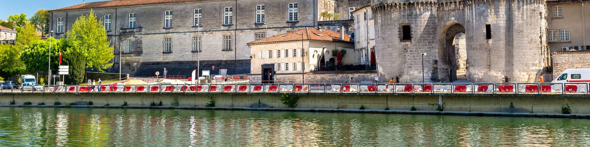 A historic building with a large arched entrance and a tower, situated by a calm river.