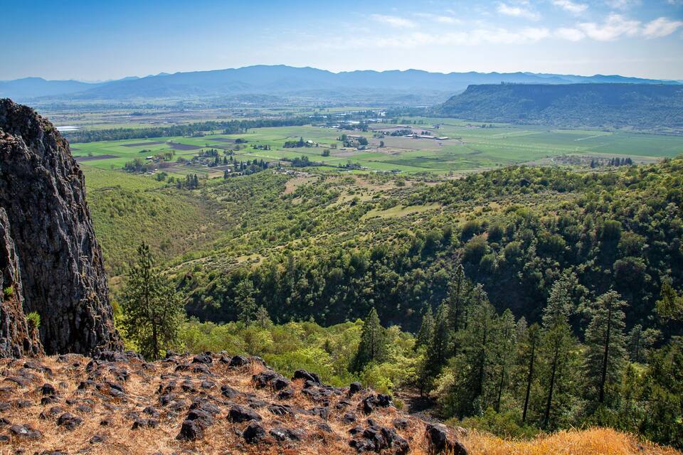 A mountainous landscape with a rocky outcrop, dense forest, and a valley with fields and small buildings.