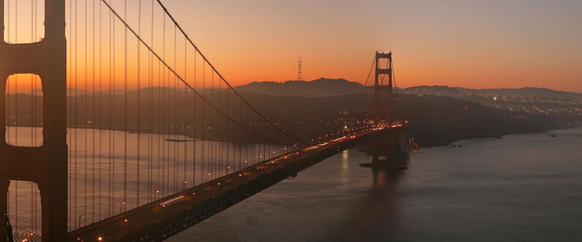 A suspension bridge at sunset with city lights in the distance.