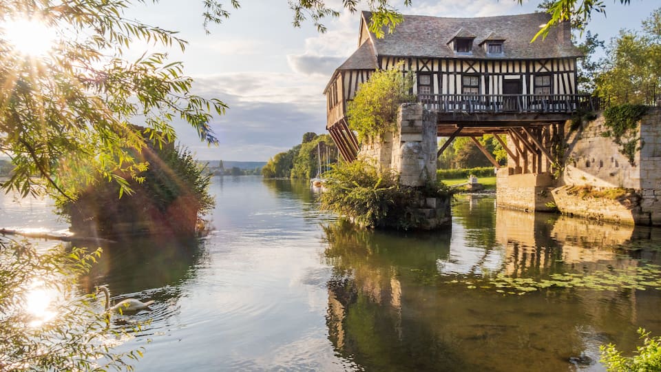 A riverside view with a traditional timber-framed house on stilts, surrounded by lush greenery and calm water.