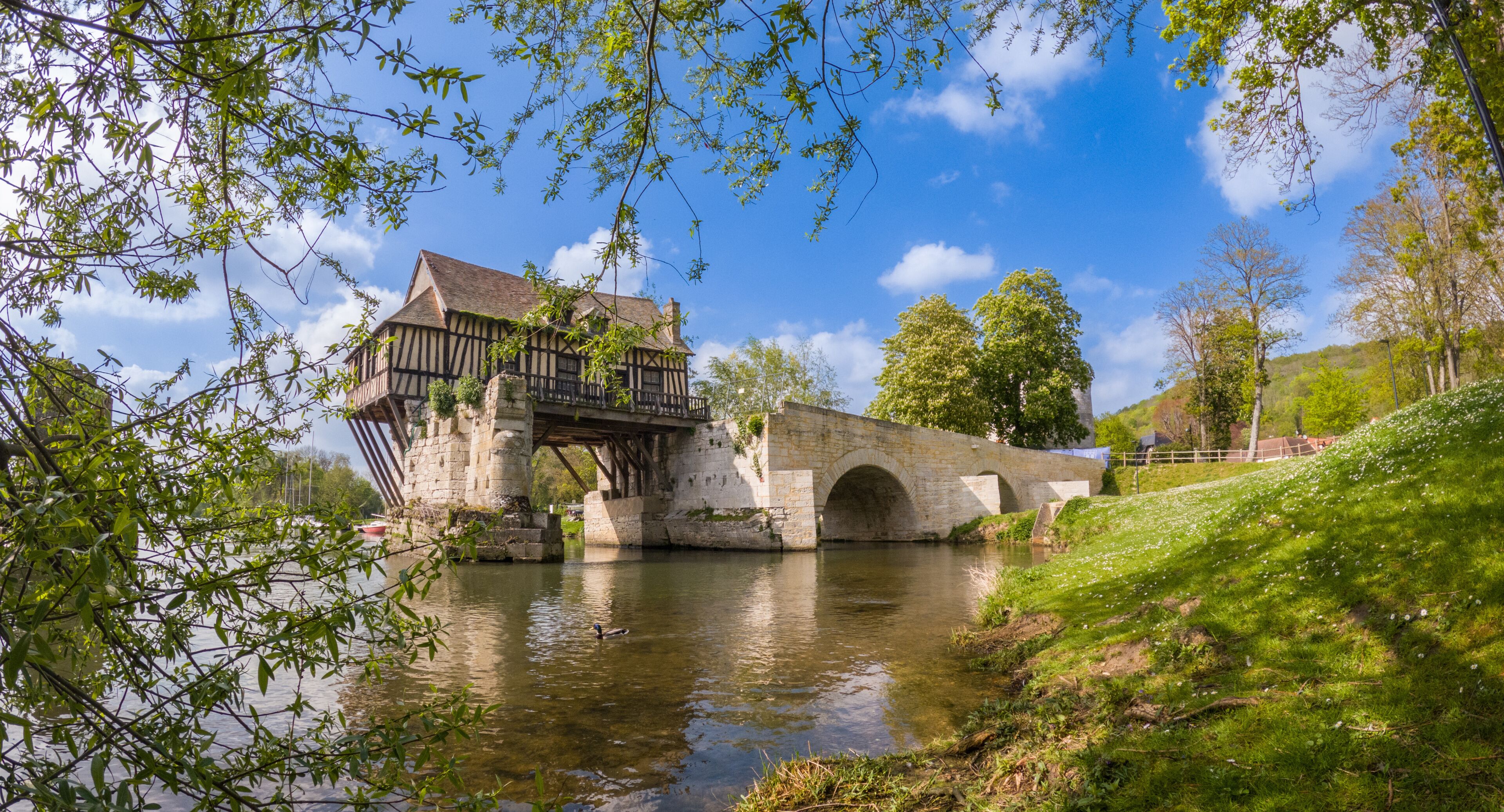 A historic stone bridge over a river, a traditional timber-framed house, and lush greenery.