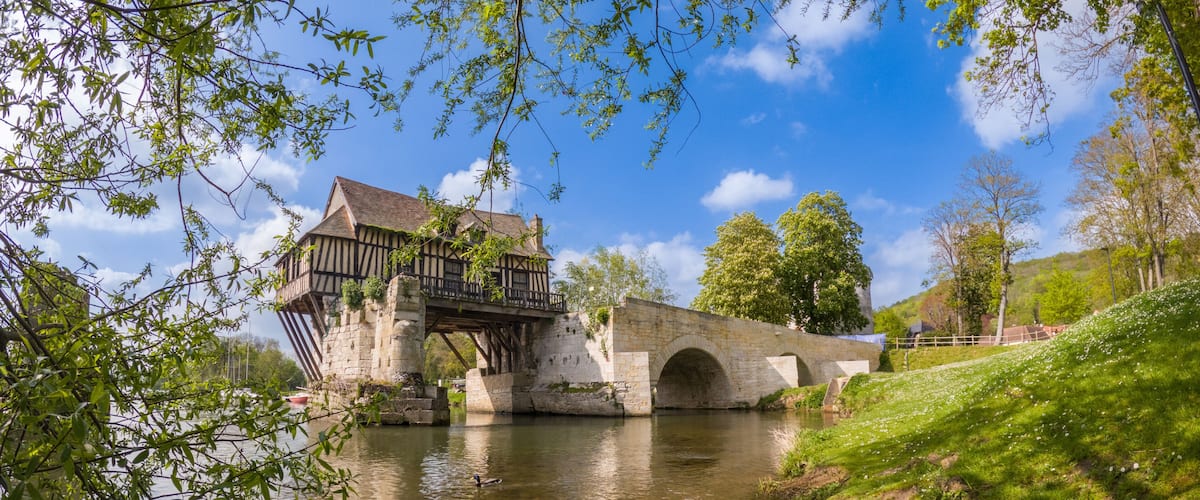 A historic stone bridge over a river, a traditional timber-framed house, and lush greenery.
