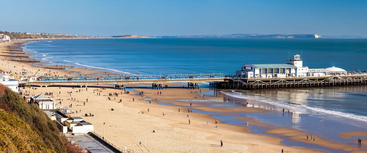 A coastal beach with a pier extending into the sea, a lighthouse, and a clear blue sky.