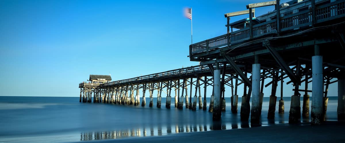 A pier extending into calm waters, a flag fluttering, and a distant building.