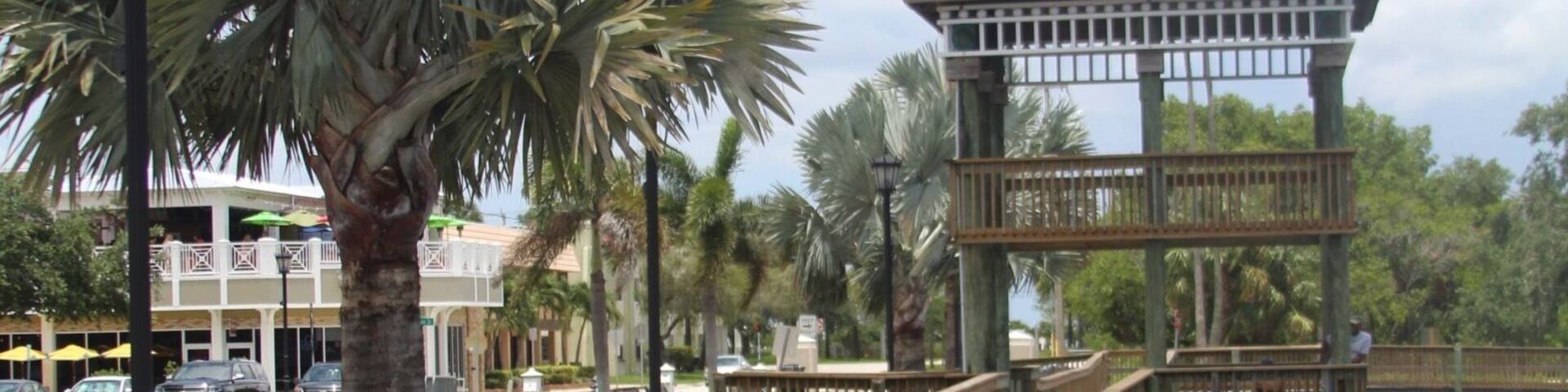 A wooden boardwalk leading to a gazebo over water, surrounded by palm trees and a building in the background.