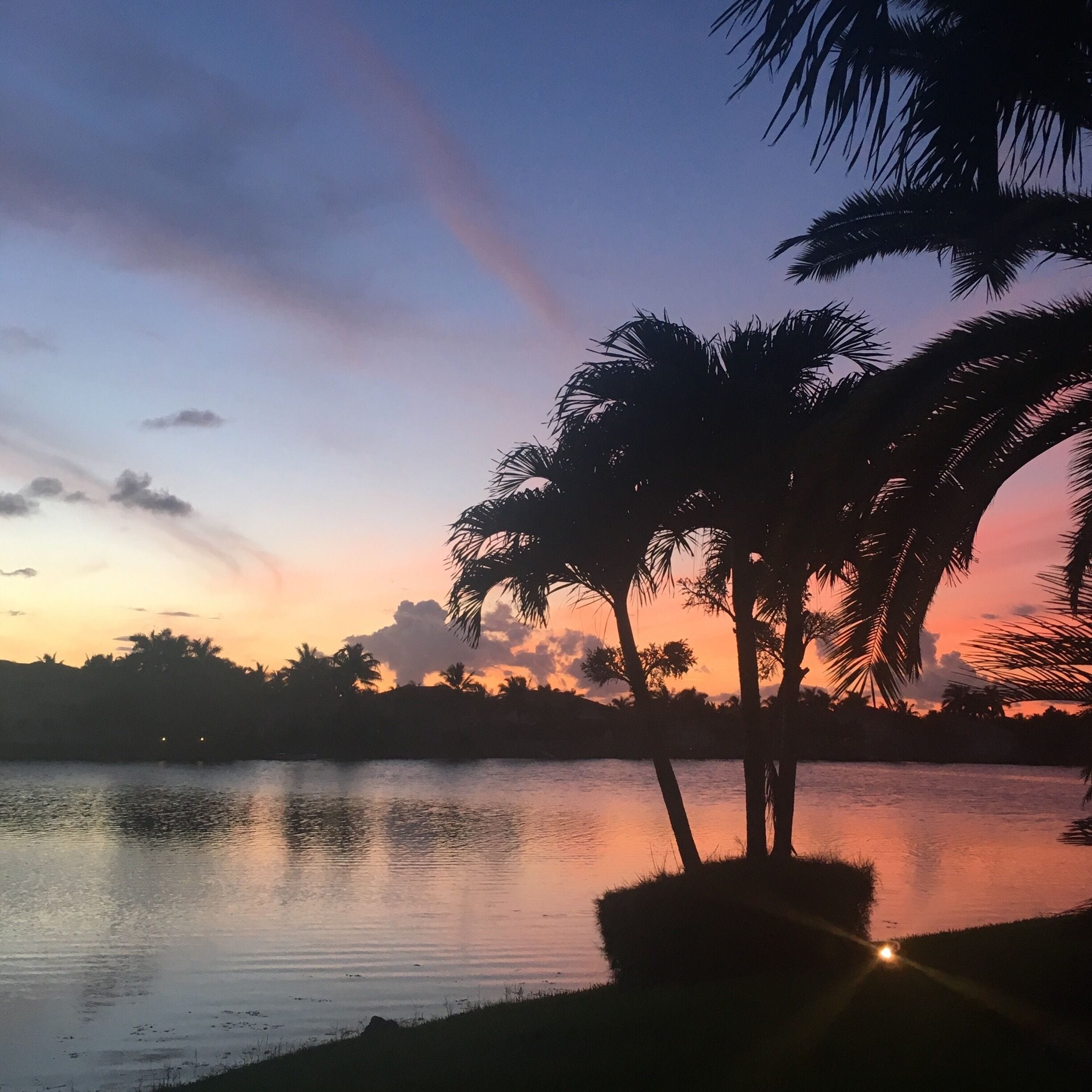 A sunset over a calm lake with silhouettes of palm trees and distant hills.