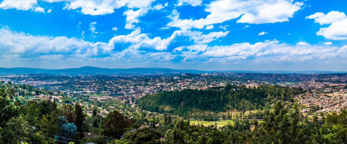 A panoramic view of a city with a dense urban area, surrounded by green hills and a clear blue sky with scattered clouds.