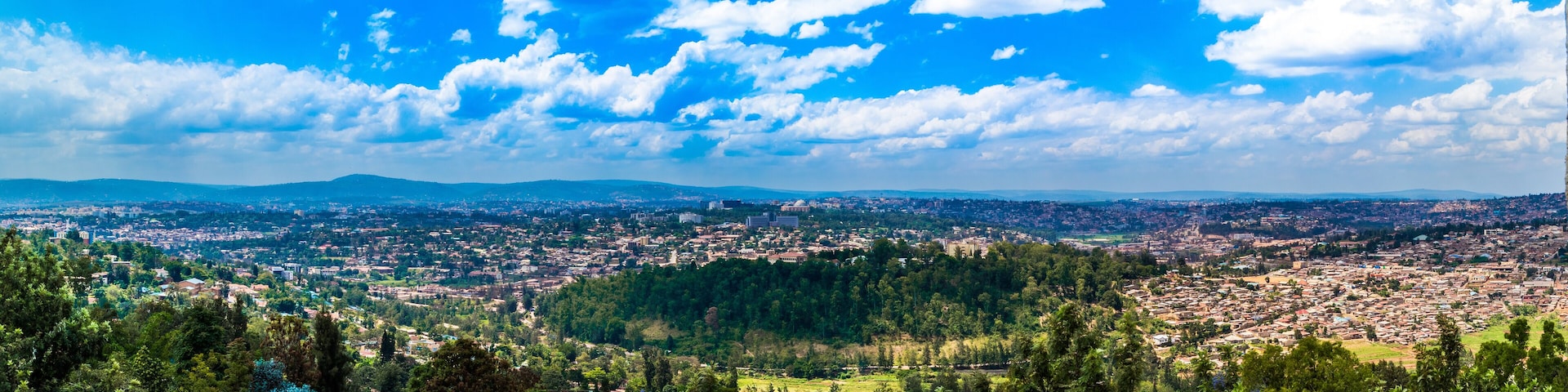 A panoramic view of a city with a dense urban area, surrounded by green hills and a clear blue sky with scattered clouds.
