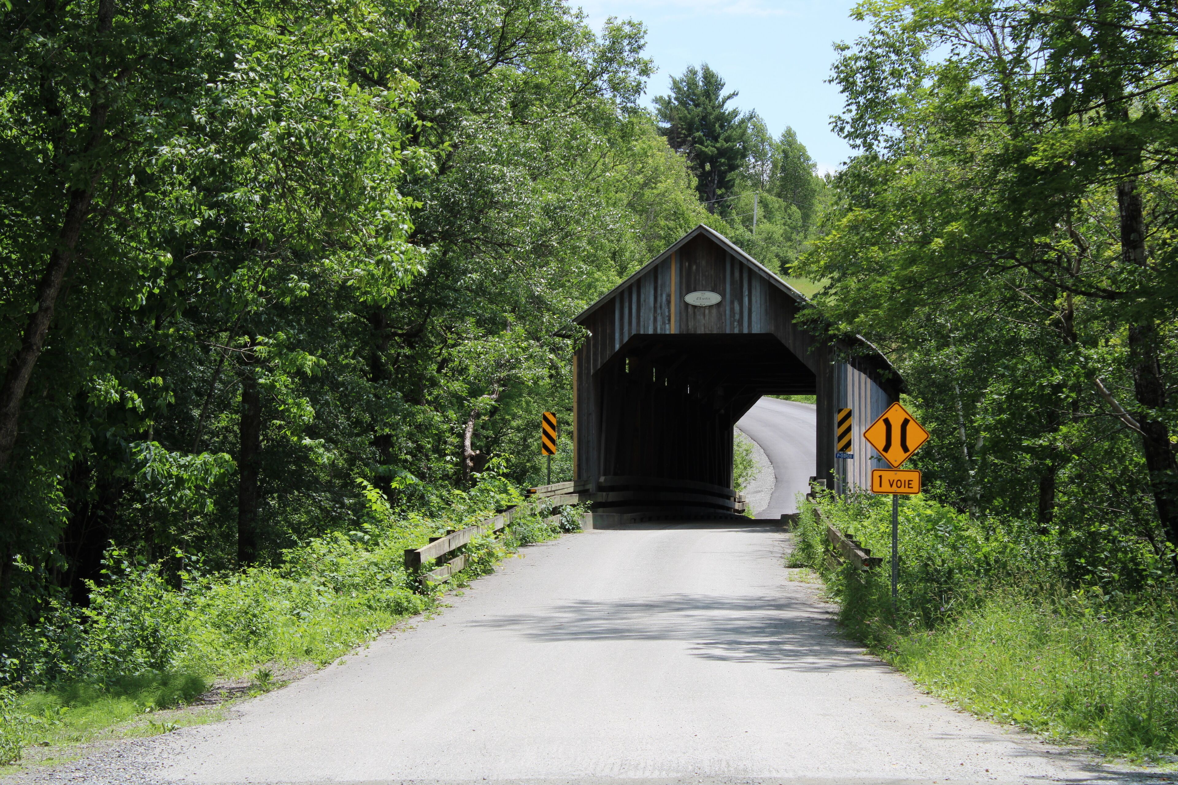 A covered bridge with a narrow road leading to it, surrounded by lush greenery.