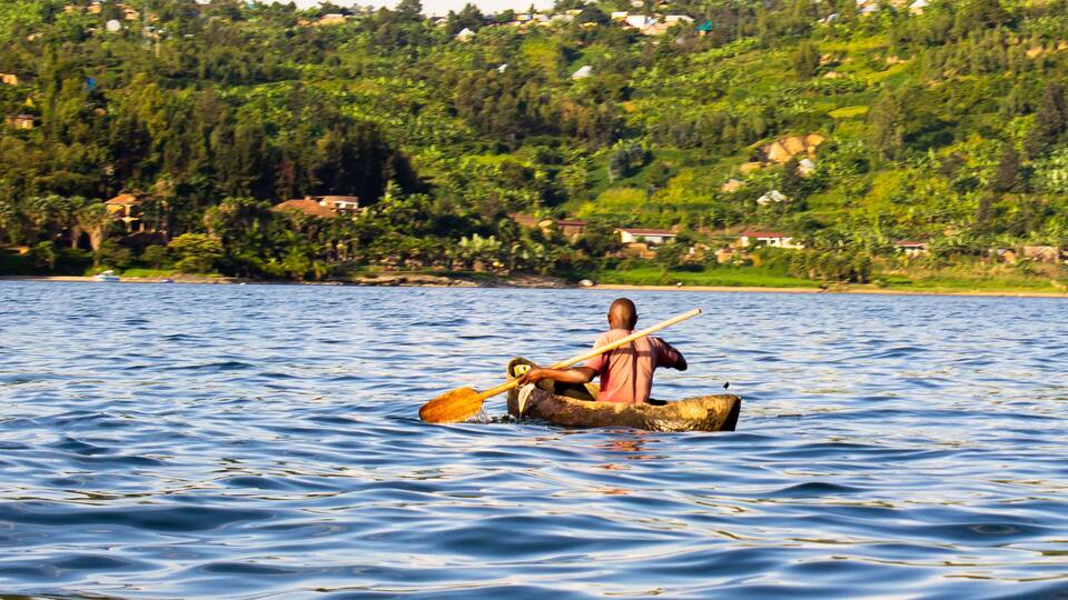 A man rowing a canoe on a calm lake with a forested hillside in the background.