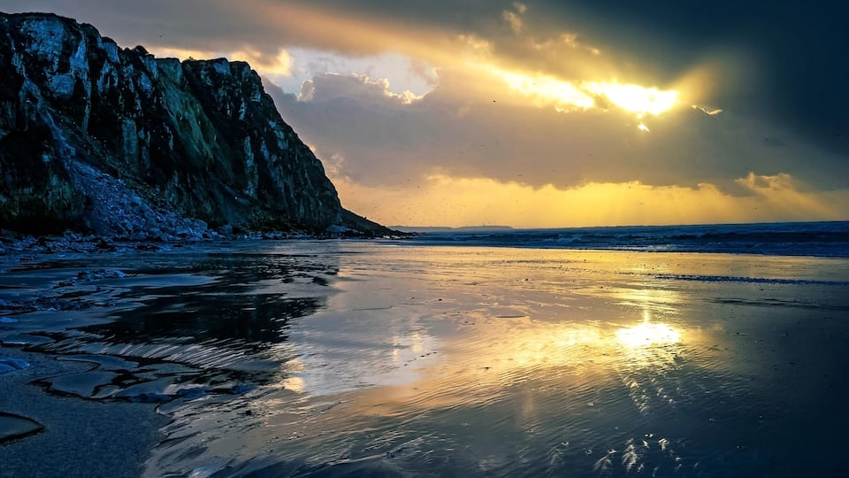 A beach with a rocky cliff, calm water reflecting the sky, and distant horizon.