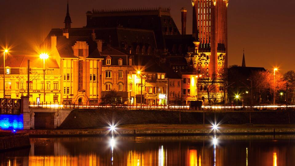 A nighttime view of a historic tower reflecting on a calm river, surrounded by illuminated buildings.