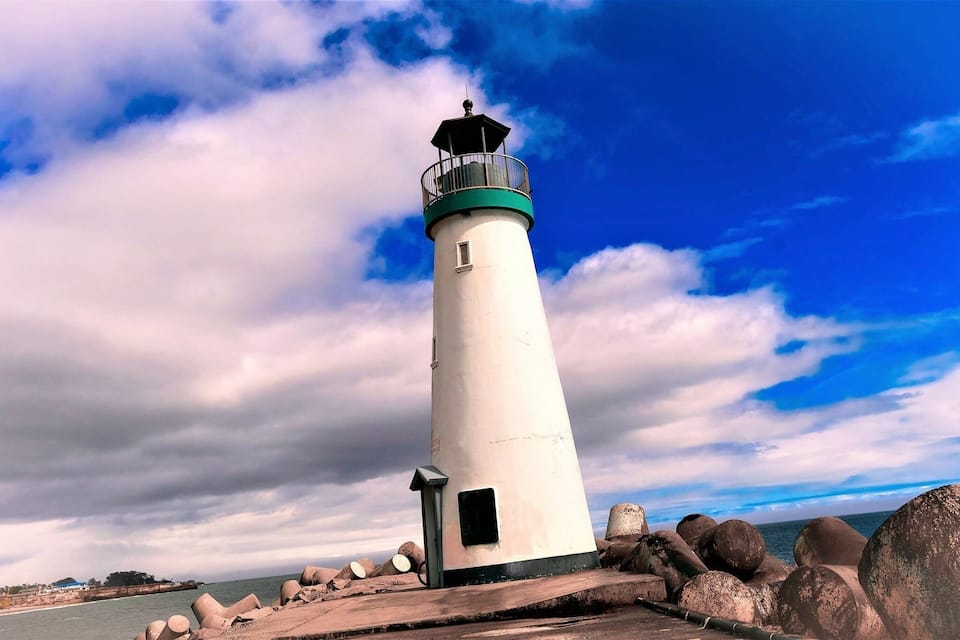 Um farol alto e branco, com a parte superior verde, rodeado por pedras grandes e um céu azul límpido.