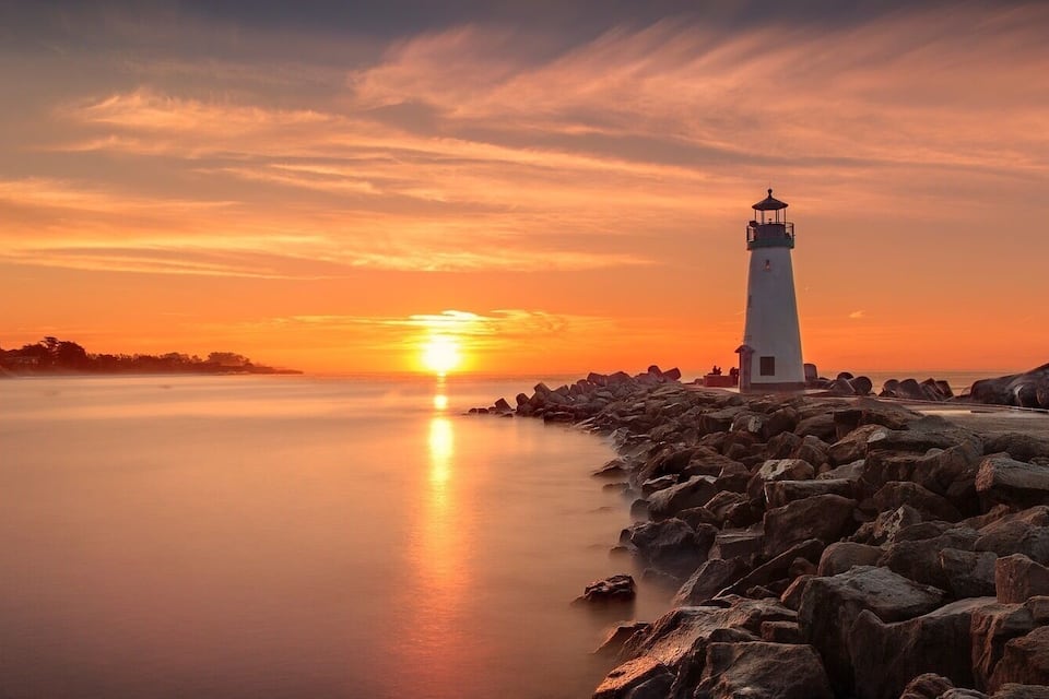 Ein Leuchtturm bei Sonnenuntergang, felsige Küstenlinie, ruhiges Wasser und klarer Himmel.