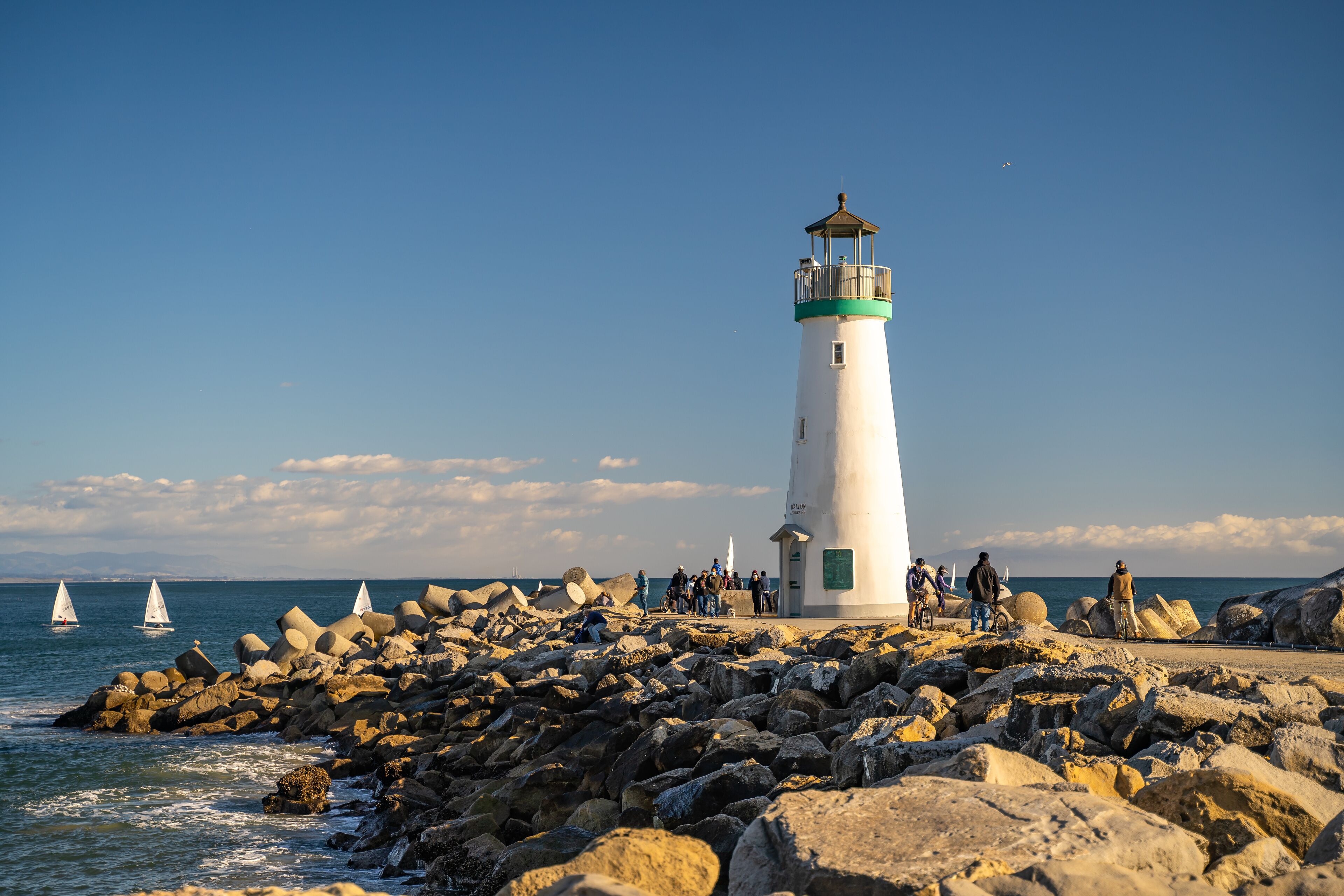 A lighthouse on a rocky shore with sailboats in the background.