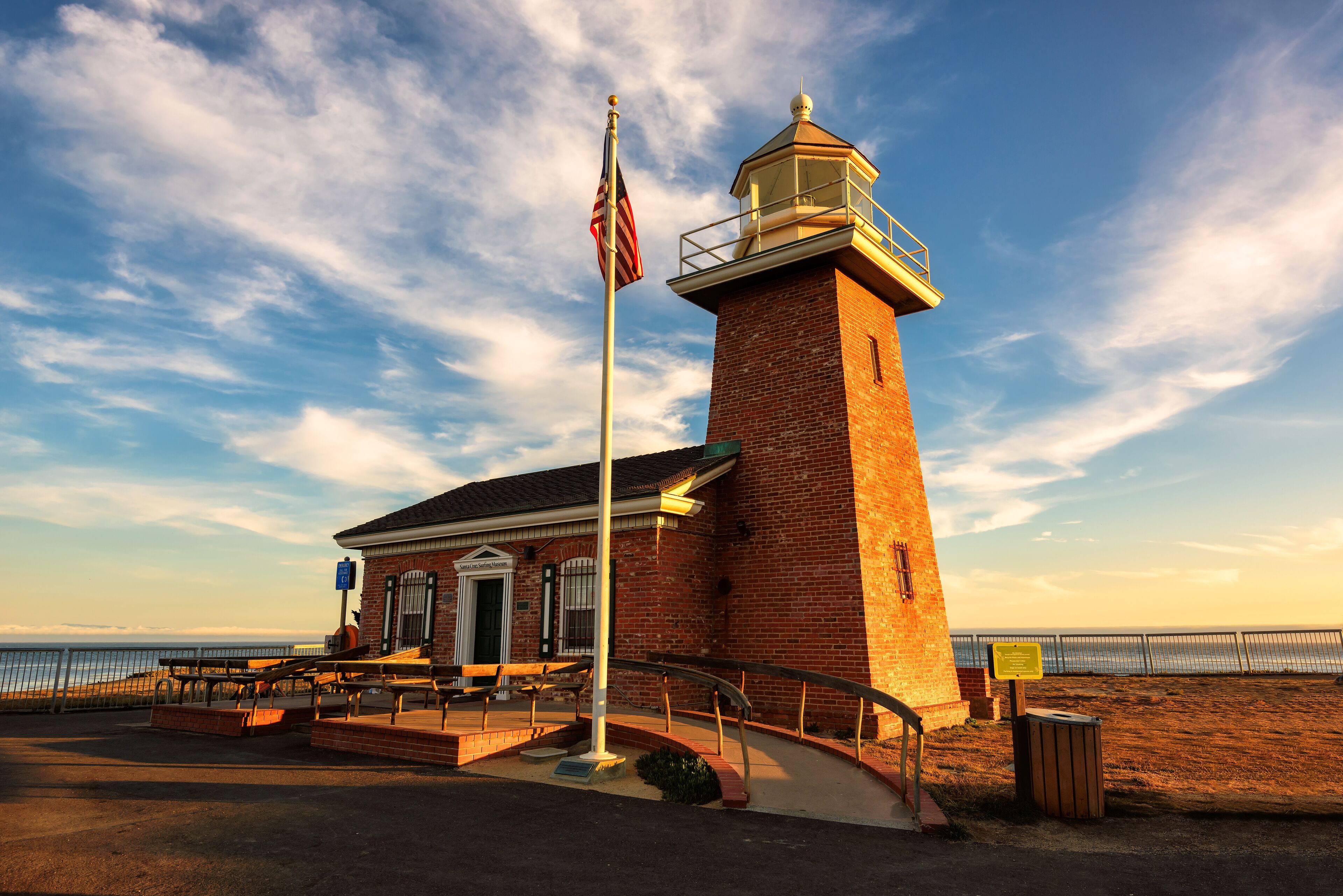 A brick lighthouse with a flagpole, a bench area, and a view of the ocean.