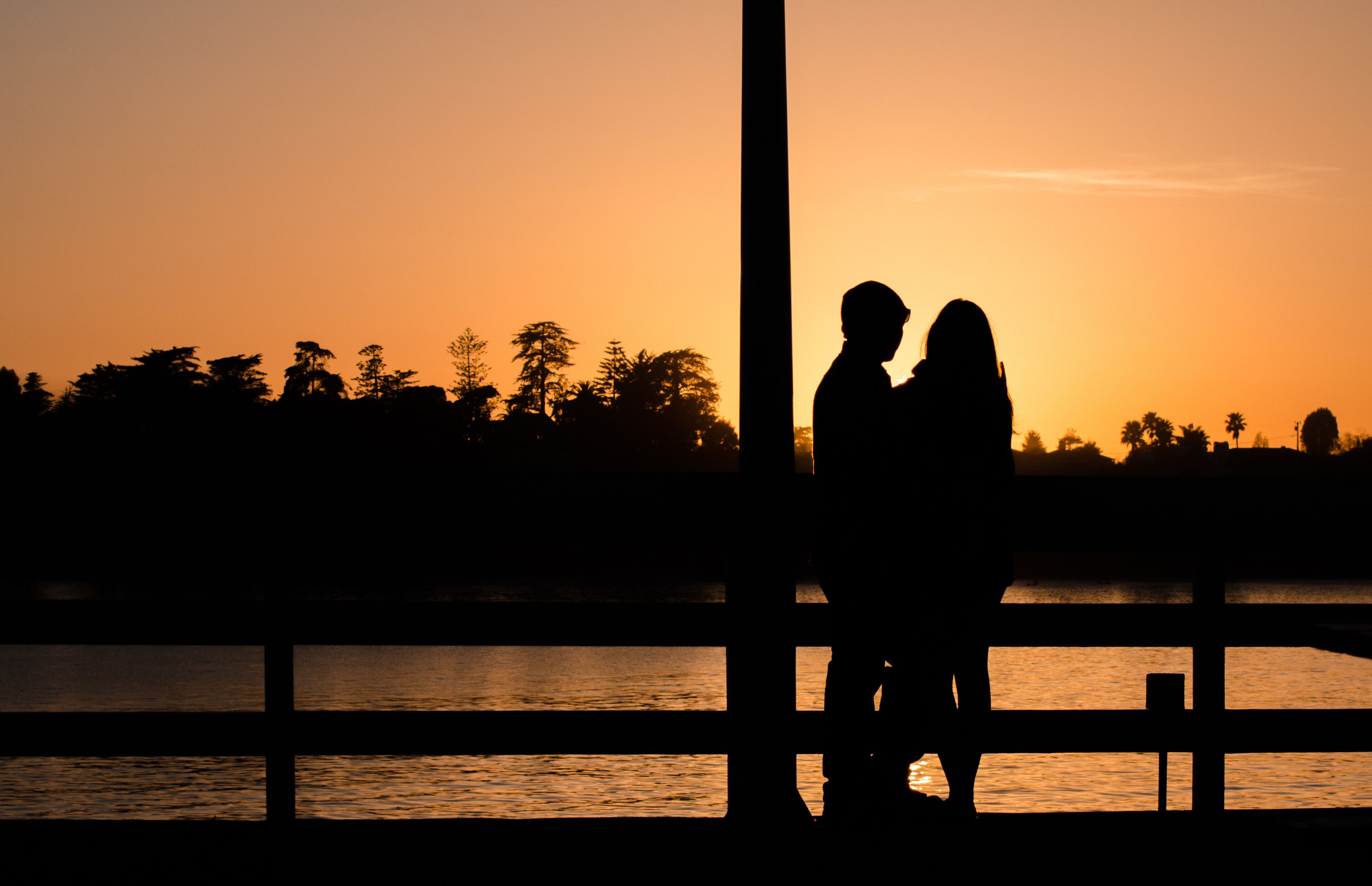 Silueta de una pareja al atardecer, con un cuerpo de agua y árboles en el fondo.