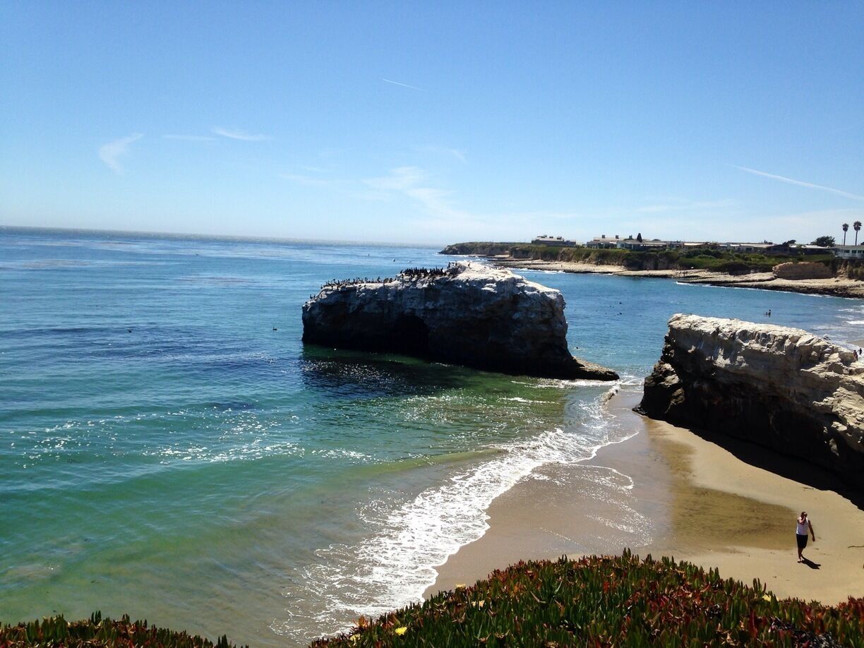 A coastal landscape with clear blue waters, a rocky outcrop, and a sandy beach.