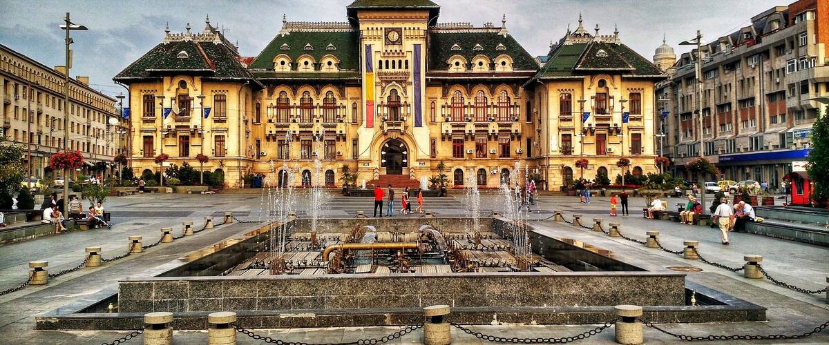A grand building with a central fountain, surrounded by a square with stone bollards and chains.