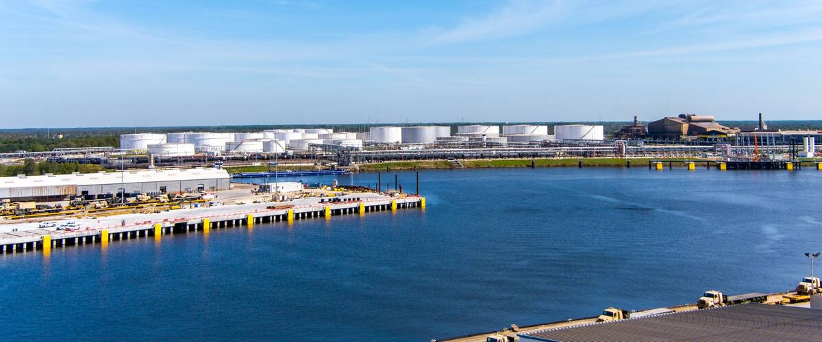 A large industrial complex with multiple storage tanks and buildings by a body of water.