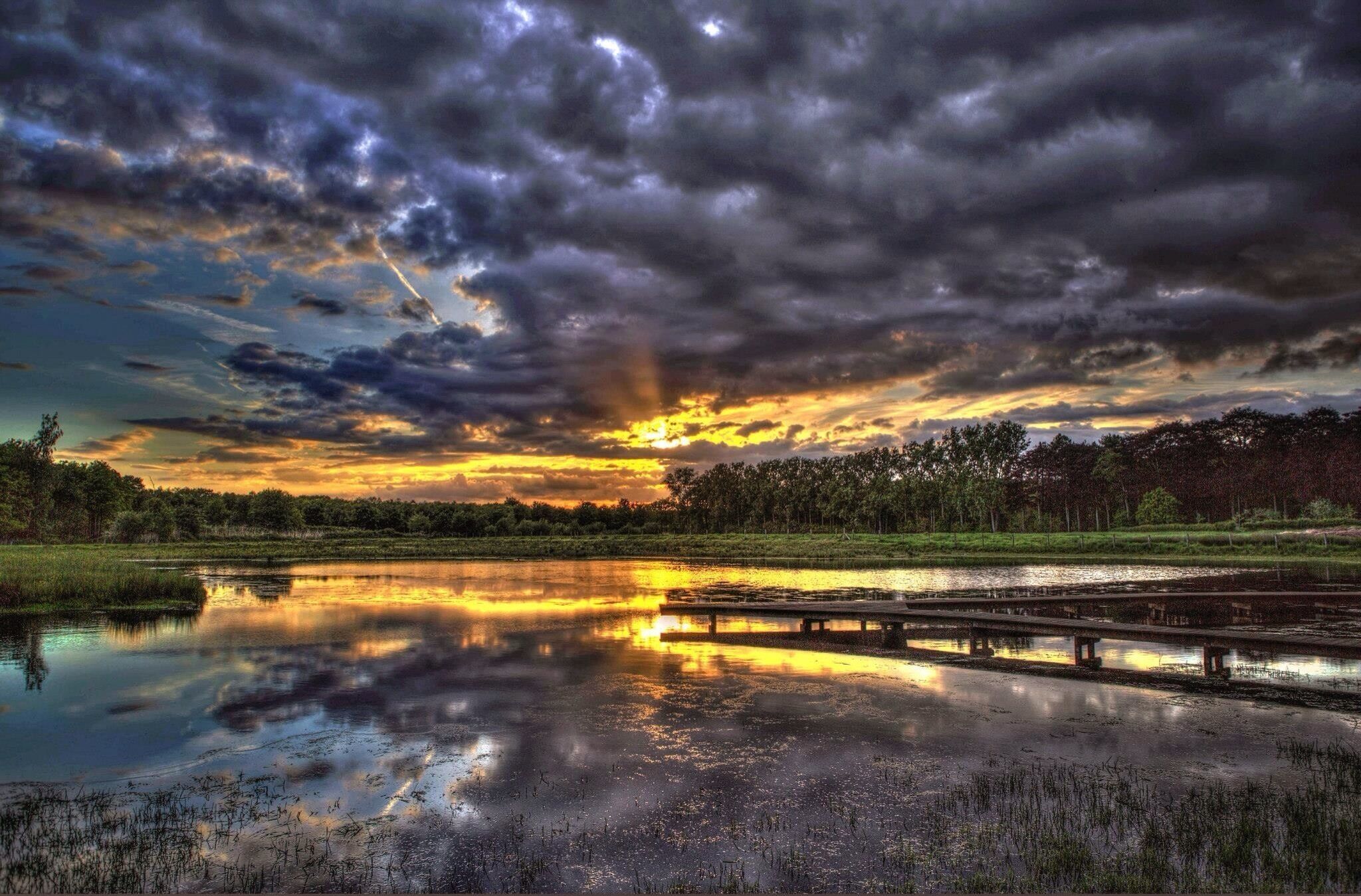 A sunset over a calm river with a wooden bridge and dense trees in the background.